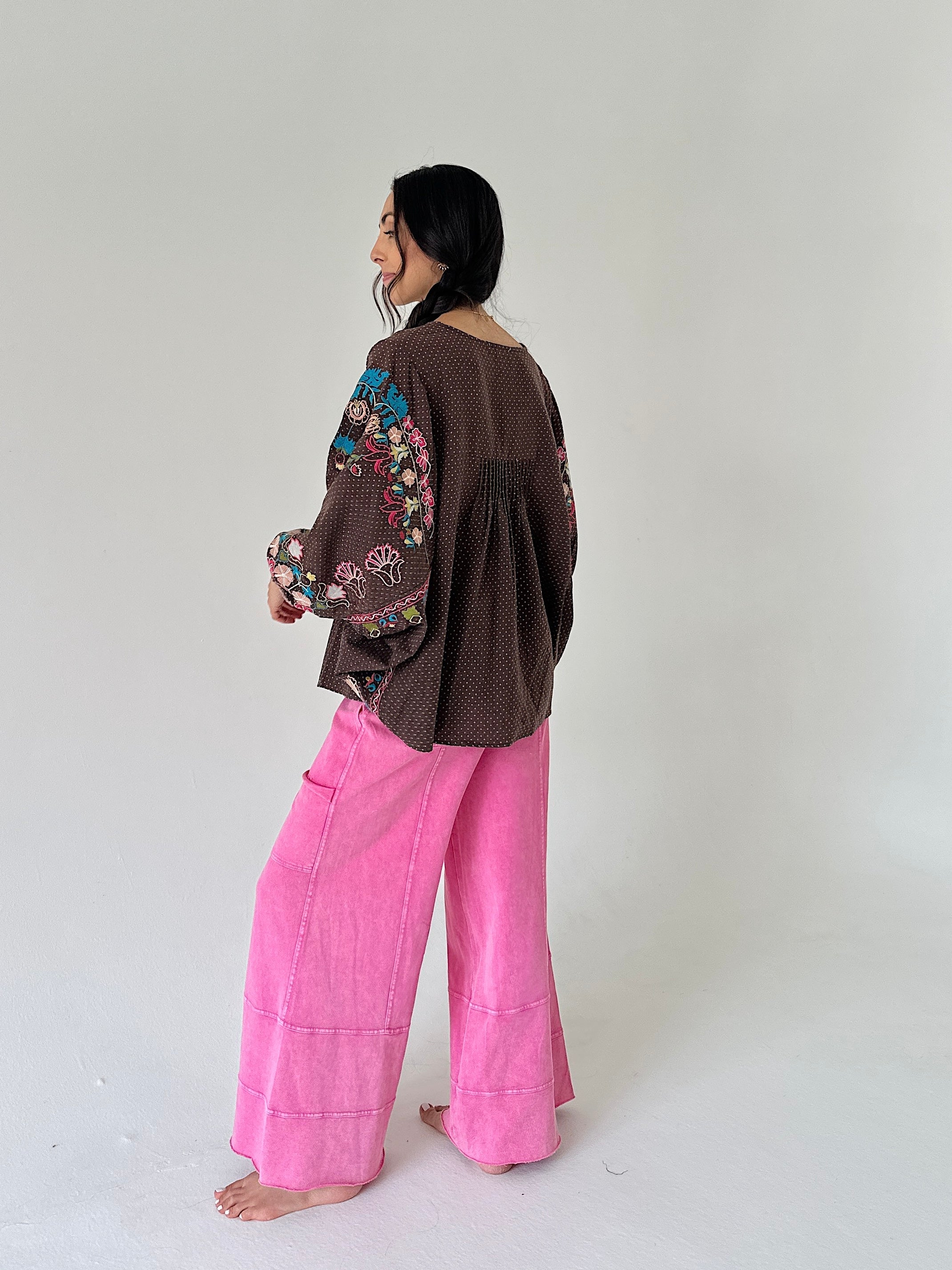 A woman stands barefoot in a studio wearing a brown long-sleeve top with colorful floral embroidery on the sleeves and bright pink wide-leg pants.