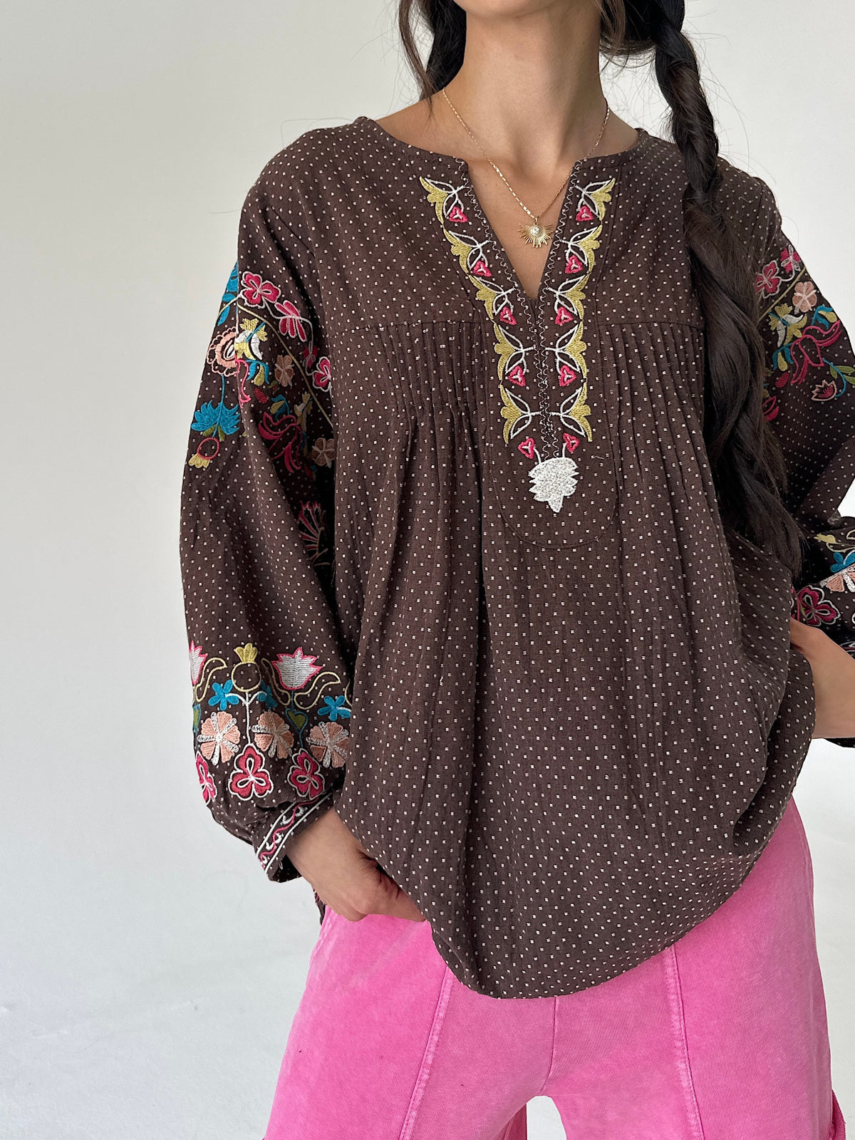 A woman wearing a brown long-sleeve blouse with a small white polka dot pattern, featuring colorful floral embroidery on the sleeves and a matching embroidered V-neckline with a white floral applique. She is also wearing bright pink pants, photographed against a plain white background.