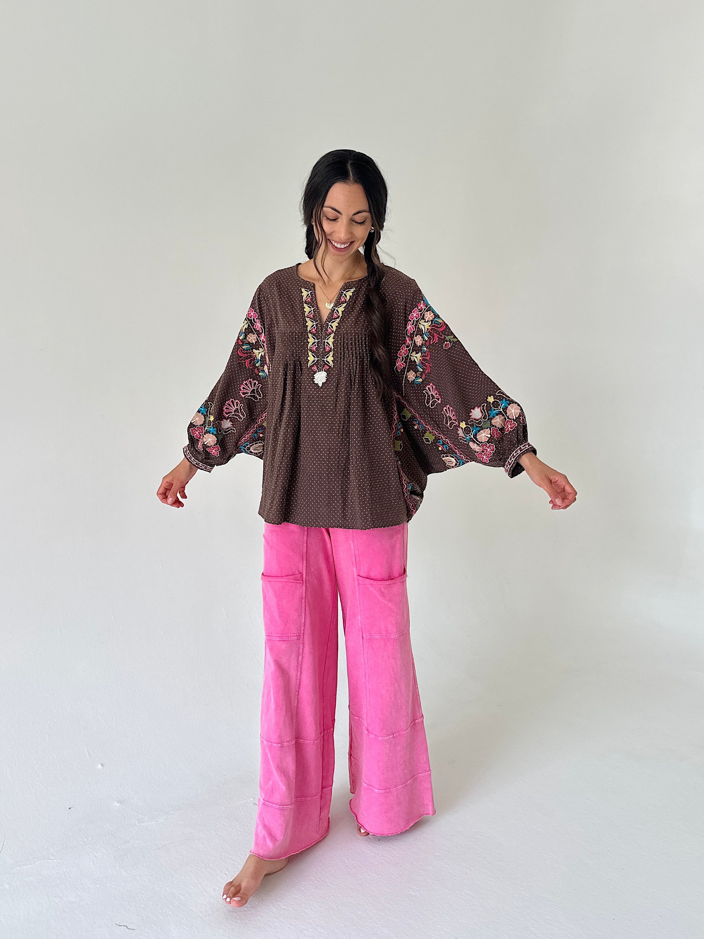 A woman stands barefoot against a plain light gray studio background, wearing a brown bohemian-style blouse with colorful floral embroidery on the sleeves and neckline, paired with bright pink wide-leg pants.