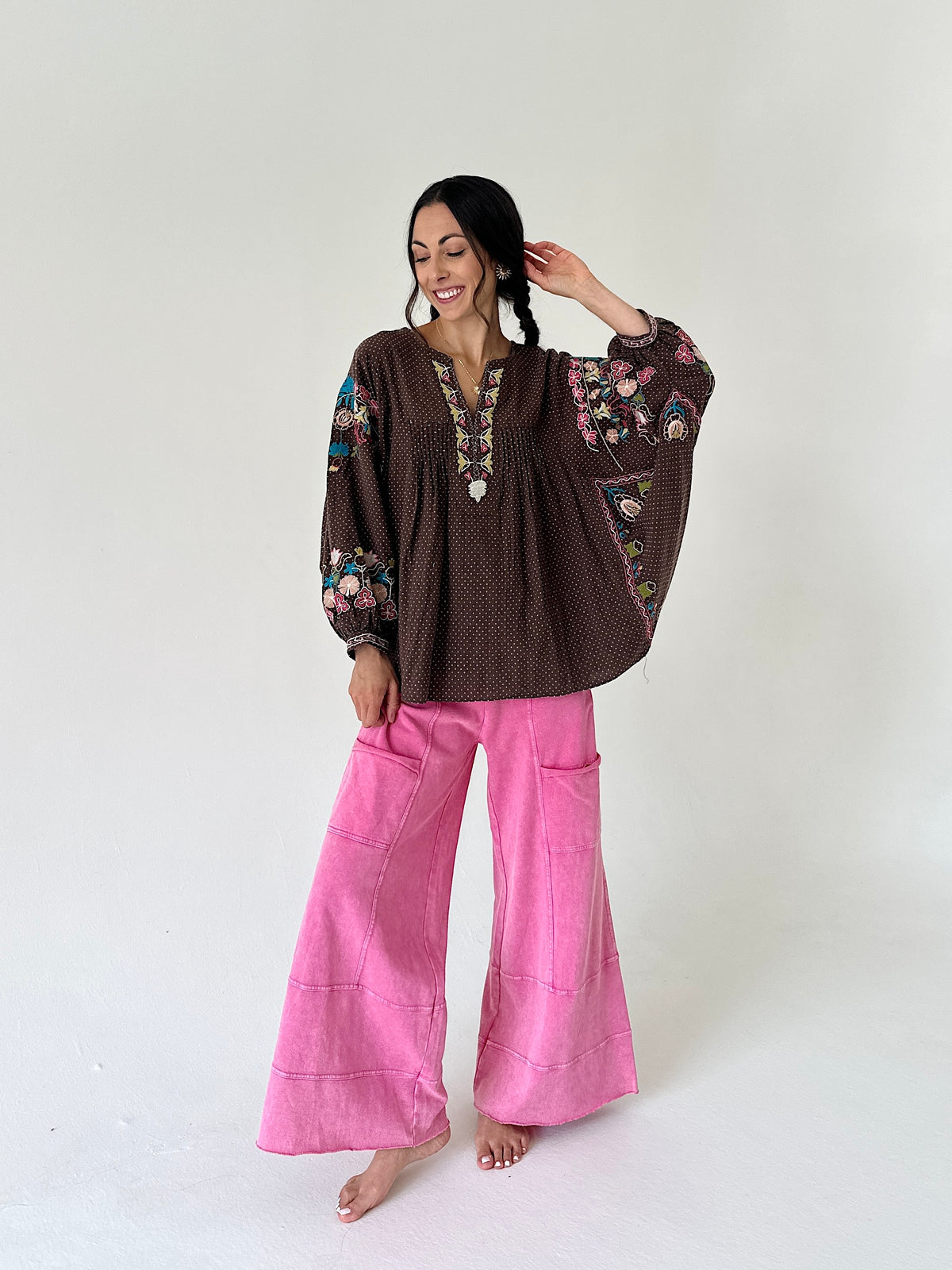 A woman stands barefoot against a plain light background wearing a brown bohemian-style embroidered blouse with colorful floral sleeve and chest details, paired with bright pink wide-leg pants.