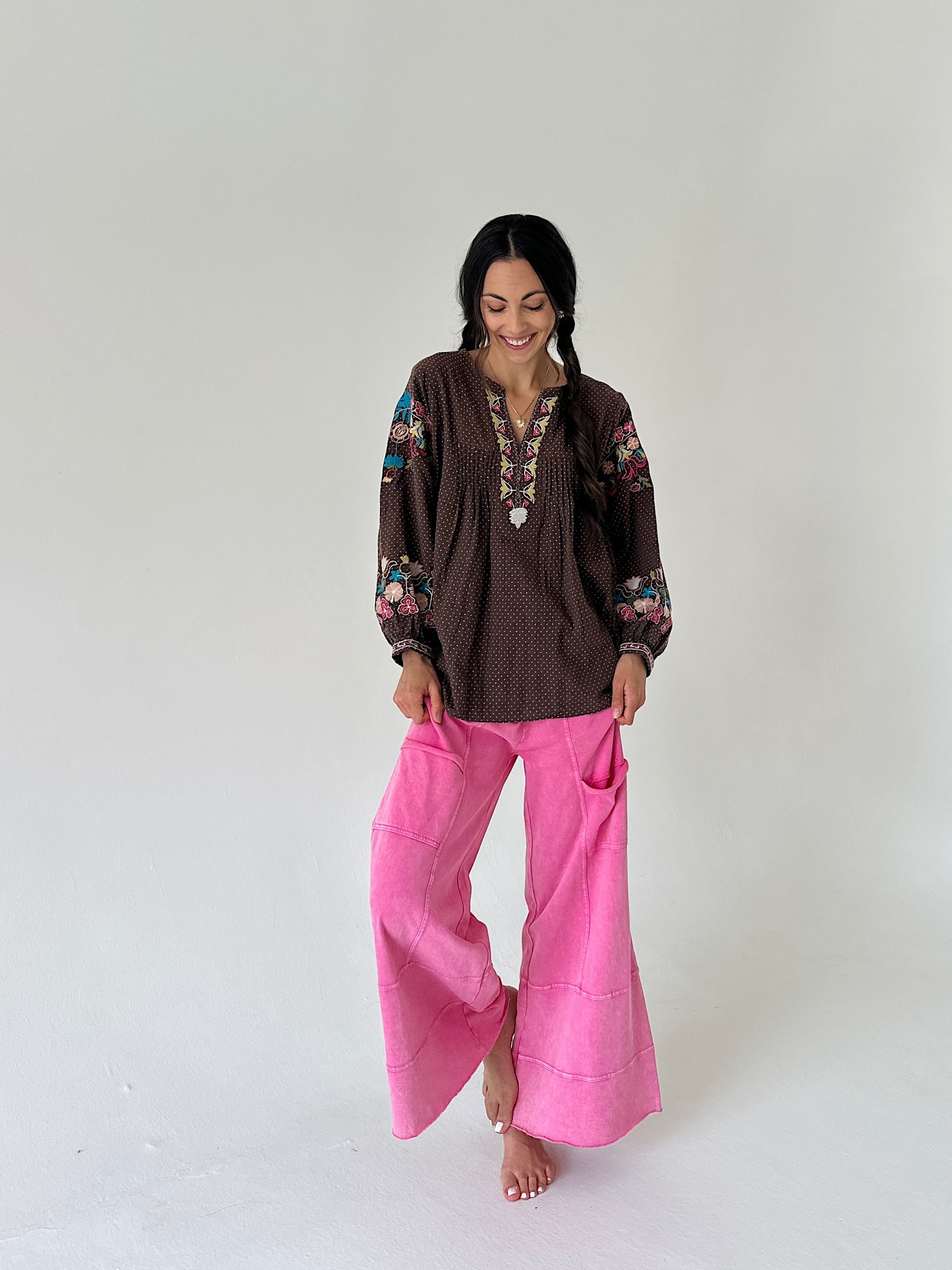 A woman modeling a brown bohemian-style embroidered blouse with colorful floral sleeve details and bright pink wide-leg pants, standing against a plain light gray studio background.