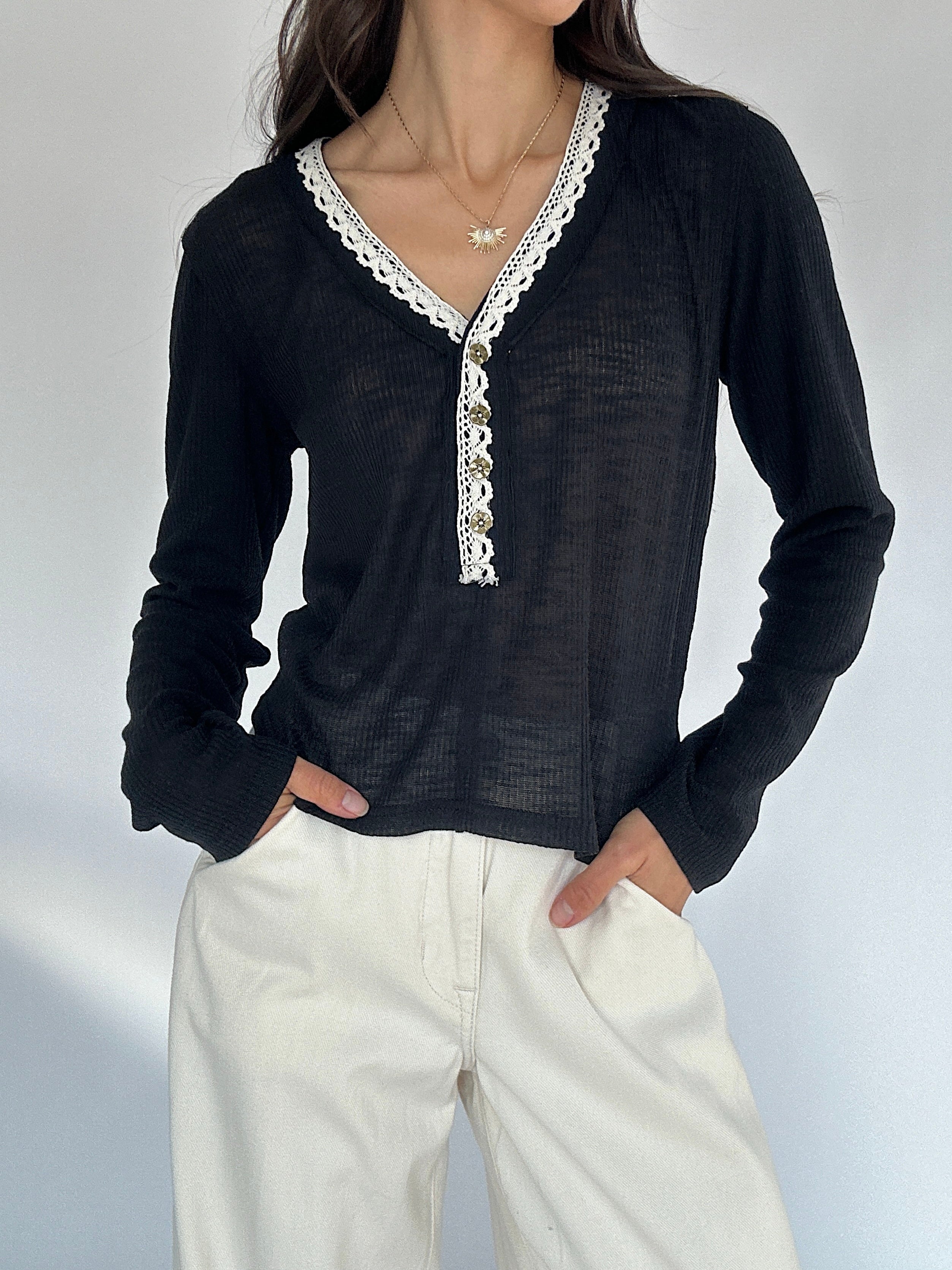 Person wearing a black long-sleeve top with white lace details and white pants on a light gray background