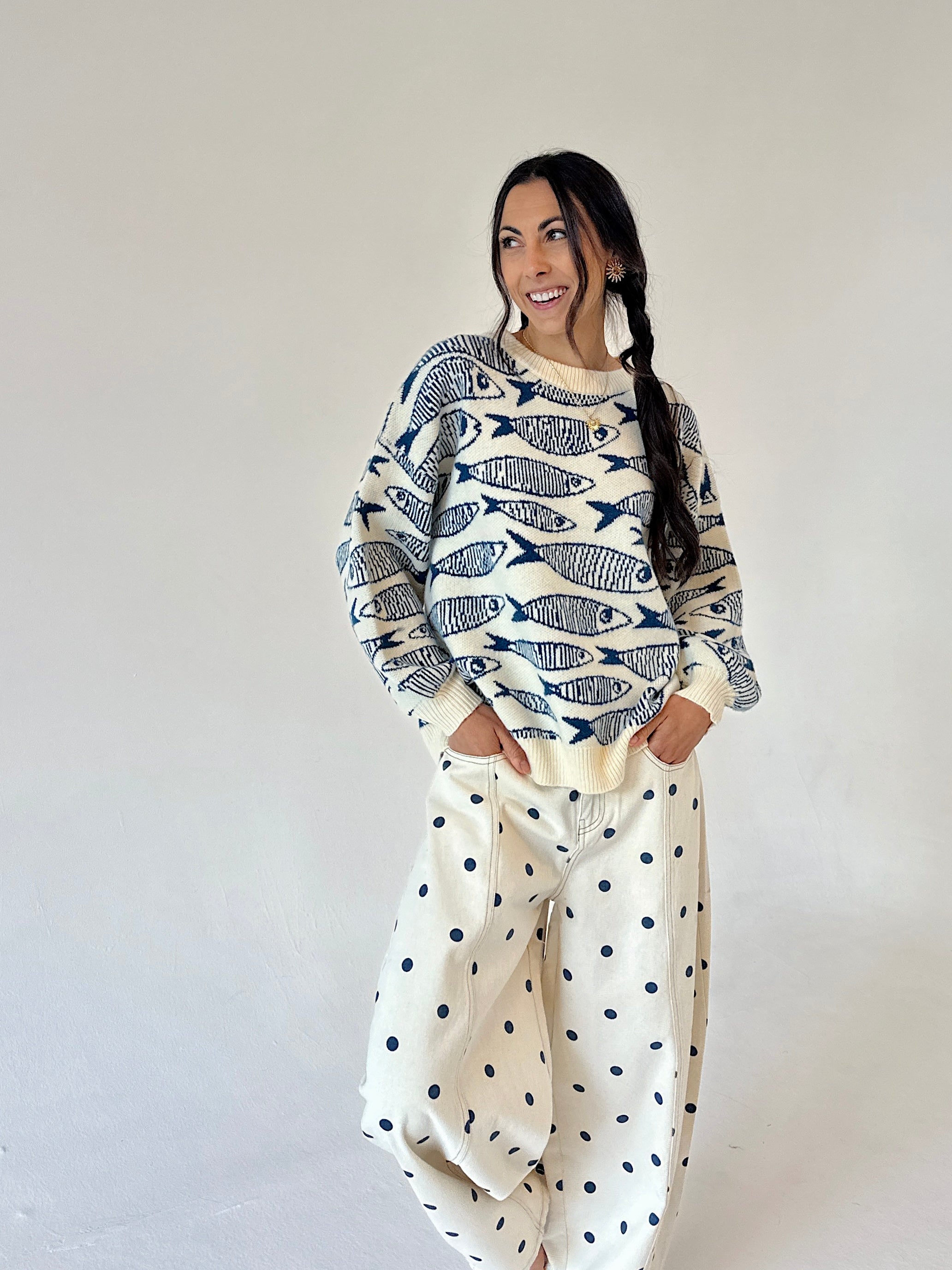 A woman modeling a cream-colored crewneck sweater with a navy blue fish pattern, paired with cream barrel-style pants with dark blue polka dots, set against a plain light gray studio background.