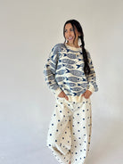 A woman modeling a cream-colored crewneck sweater with a navy blue fish pattern, paired with cream barrel-style pants with dark blue polka dots, set against a plain light gray studio background.