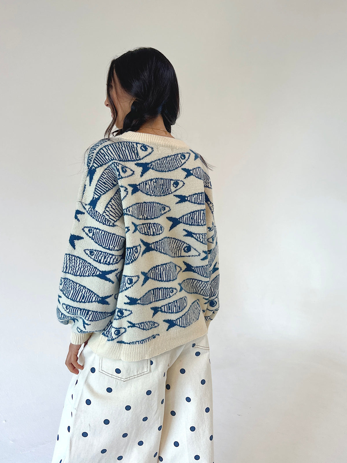 A woman shown from behind wearing a cream-colored knit sweater with a blue fish pattern and cream wide-leg pants with blue polka dots, set against a plain off-white wall.