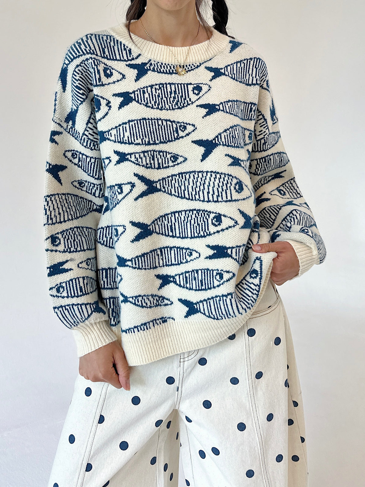 A person wearing a cream knit sweater with a repeating blue fish pattern, paired with cream pants featuring dark blue polka dots, against a plain light background.