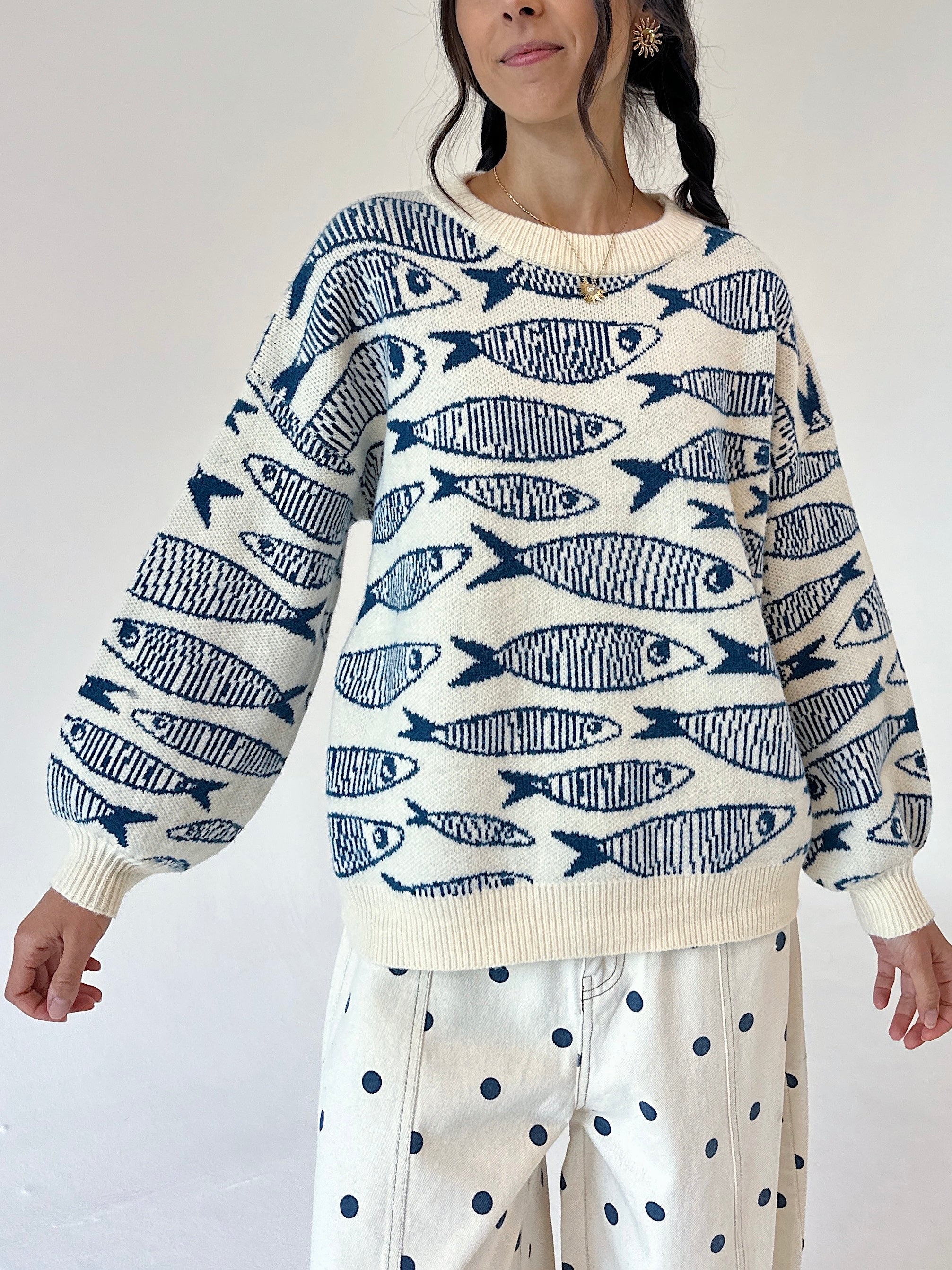 A woman wearing a cream-colored knit sweater with a navy blue fish pattern, paired with cream pants featuring navy blue polka dots, against a plain white background.