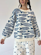 A woman wearing a cream-colored knit sweater with a navy blue fish pattern, paired with cream pants featuring navy blue polka dots, against a plain white background.