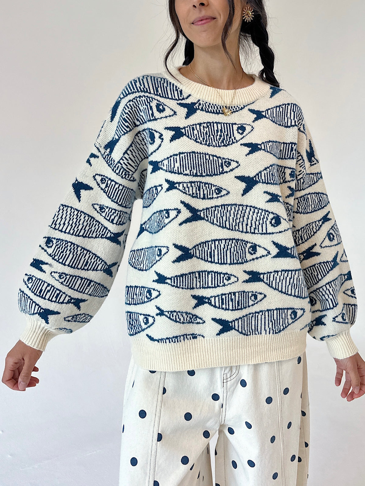 A woman wearing a cream-colored knit sweater with a navy blue fish pattern, paired with cream pants featuring navy blue polka dots, against a plain white background.