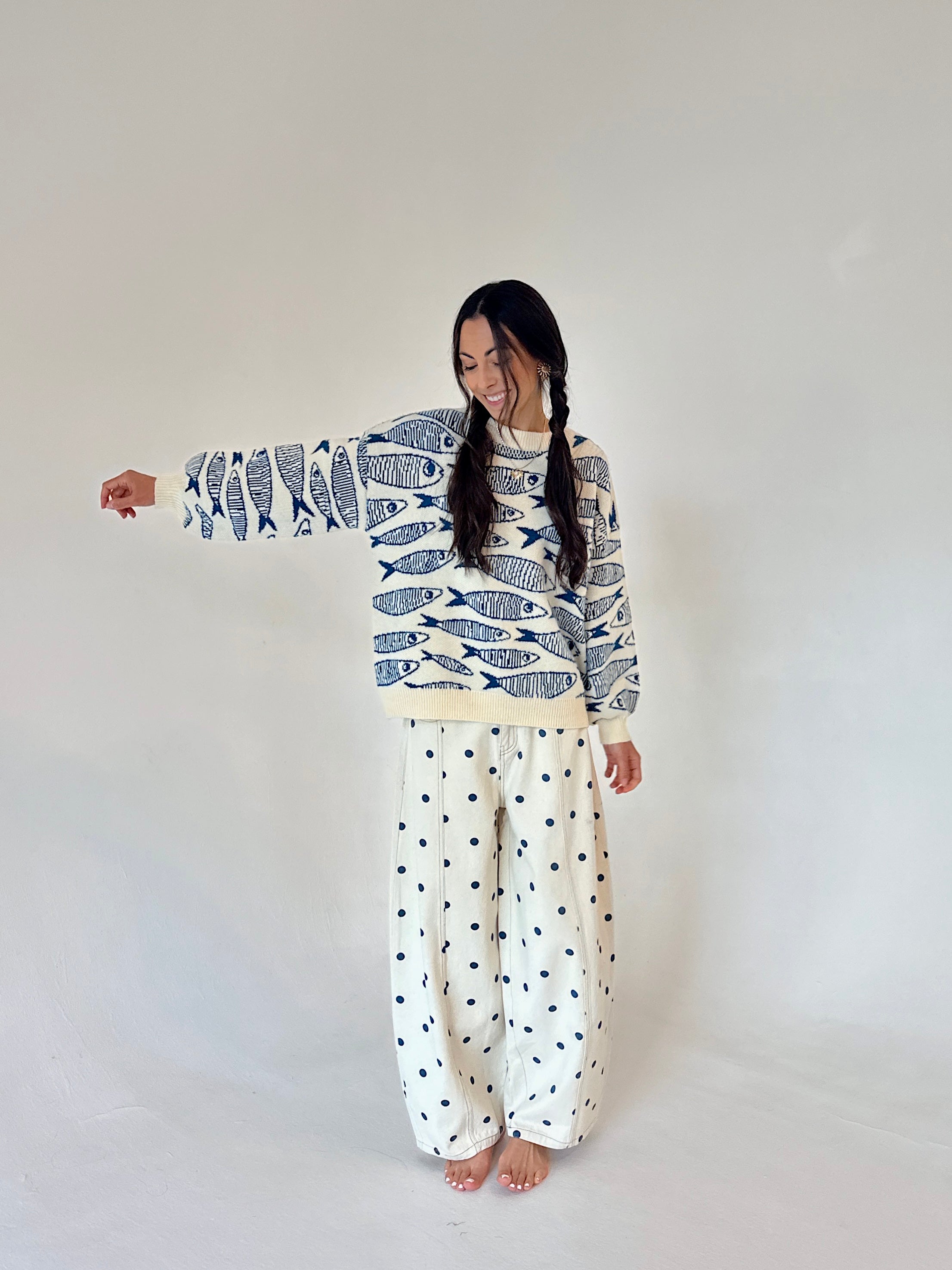 A woman modeling a long-sleeve sweater with a blue and white fish pattern and wide-leg pants with a small blue polka dot pattern on an off-white background, set against a plain light gray studio backdrop.