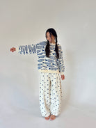 A woman modeling a long-sleeve sweater with a blue and white fish pattern and wide-leg pants with a small blue polka dot pattern on an off-white background, set against a plain light gray studio backdrop.