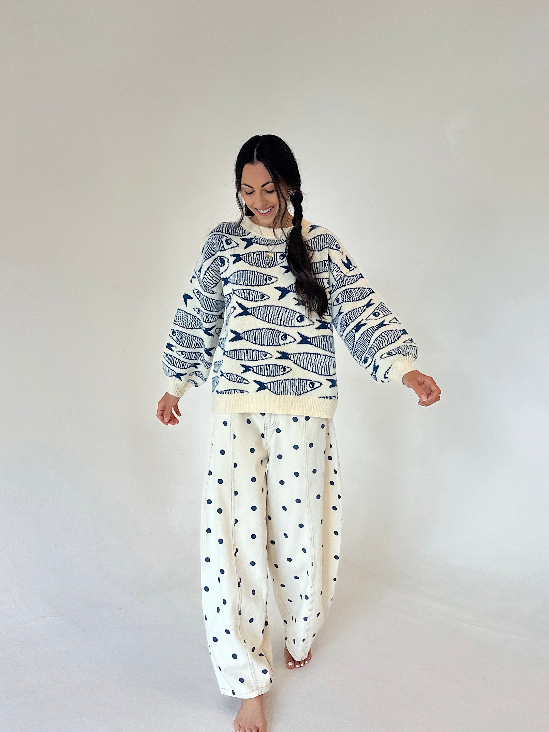 A woman modeling a cream sweater with a navy fish pattern and cream pants with navy polka dots, set against a plain light gray studio background.