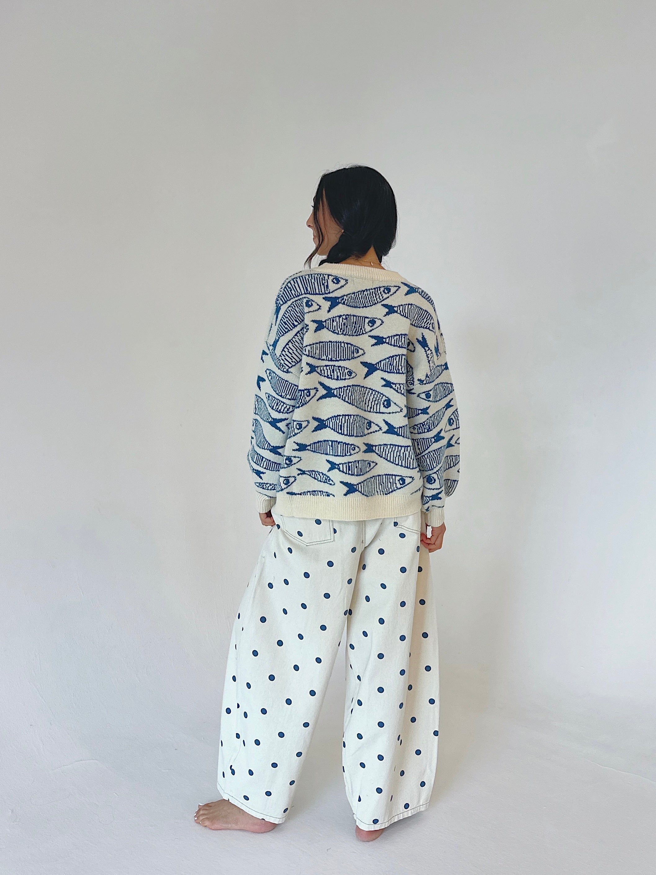 A person standing barefoot against a plain light gray wall, viewed from the back. They are wearing a cream knit sweater covered in a blue fish pattern and matching cream wide-leg pants with blue polka dots.