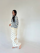 A woman stands barefoot against a plain off-white studio background, wearing a cream and navy fish patterned sweater with cream pants featuring navy polka dots.