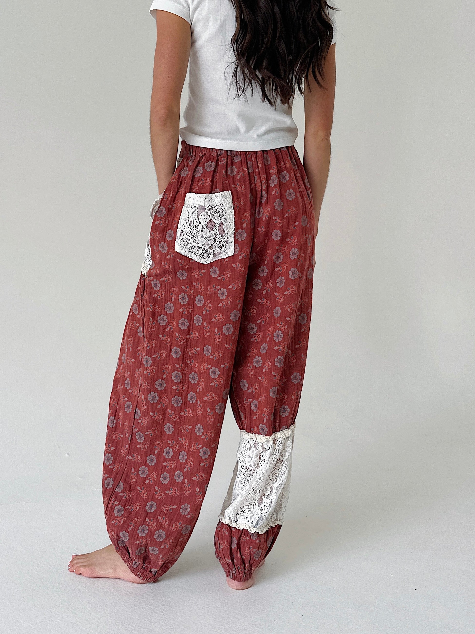 A person shown from the back wearing loose, patterned harem-style pants in a muted red-brown tone with white lace pocket and lower-leg panels, paired with a white top against a plain light gray studio background.