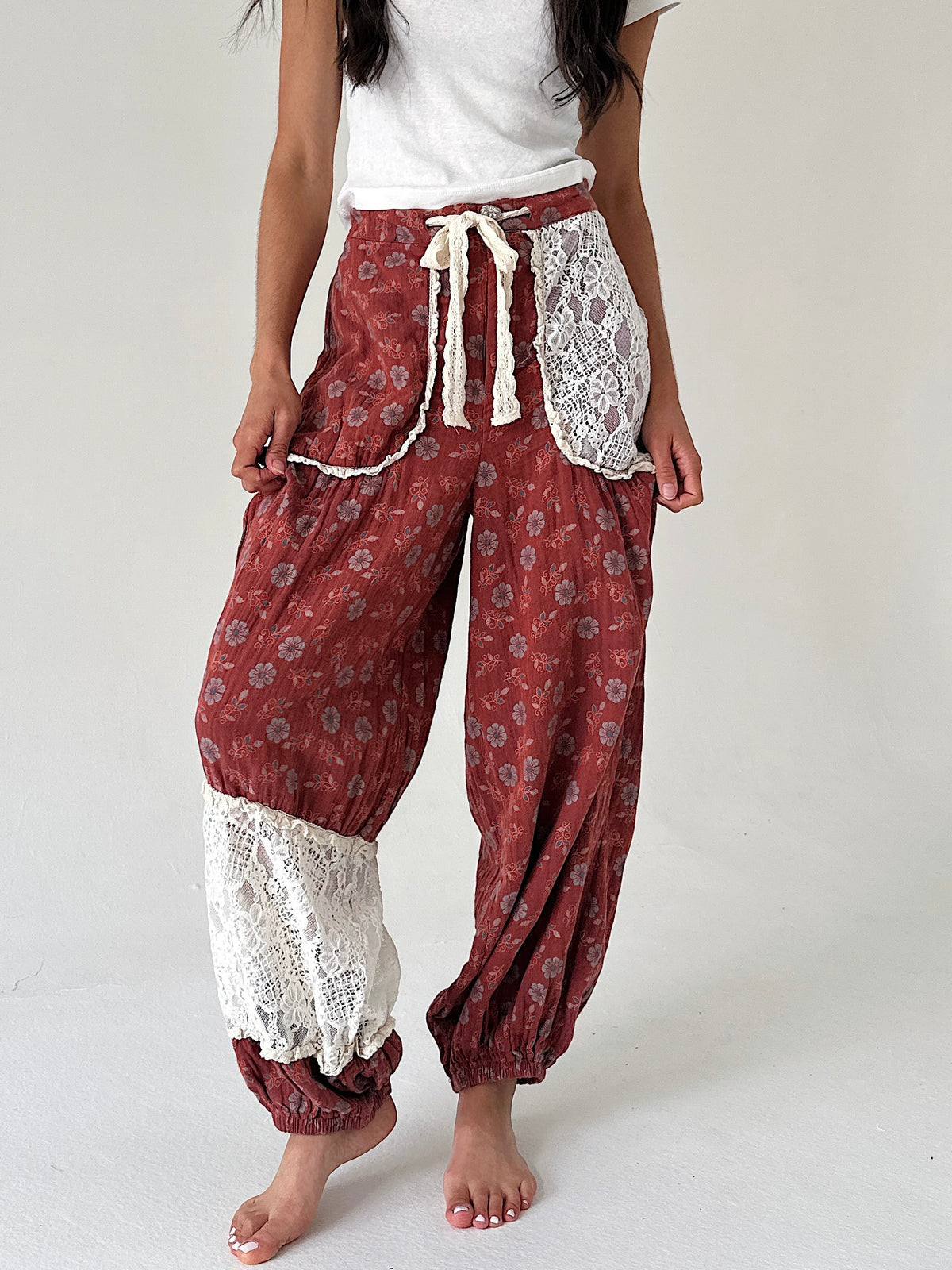 A person wearing loose, draped harem-style pants in a rust red floral print with white lace trim and a cream drawstring, paired with a white top, photographed against a plain light background.