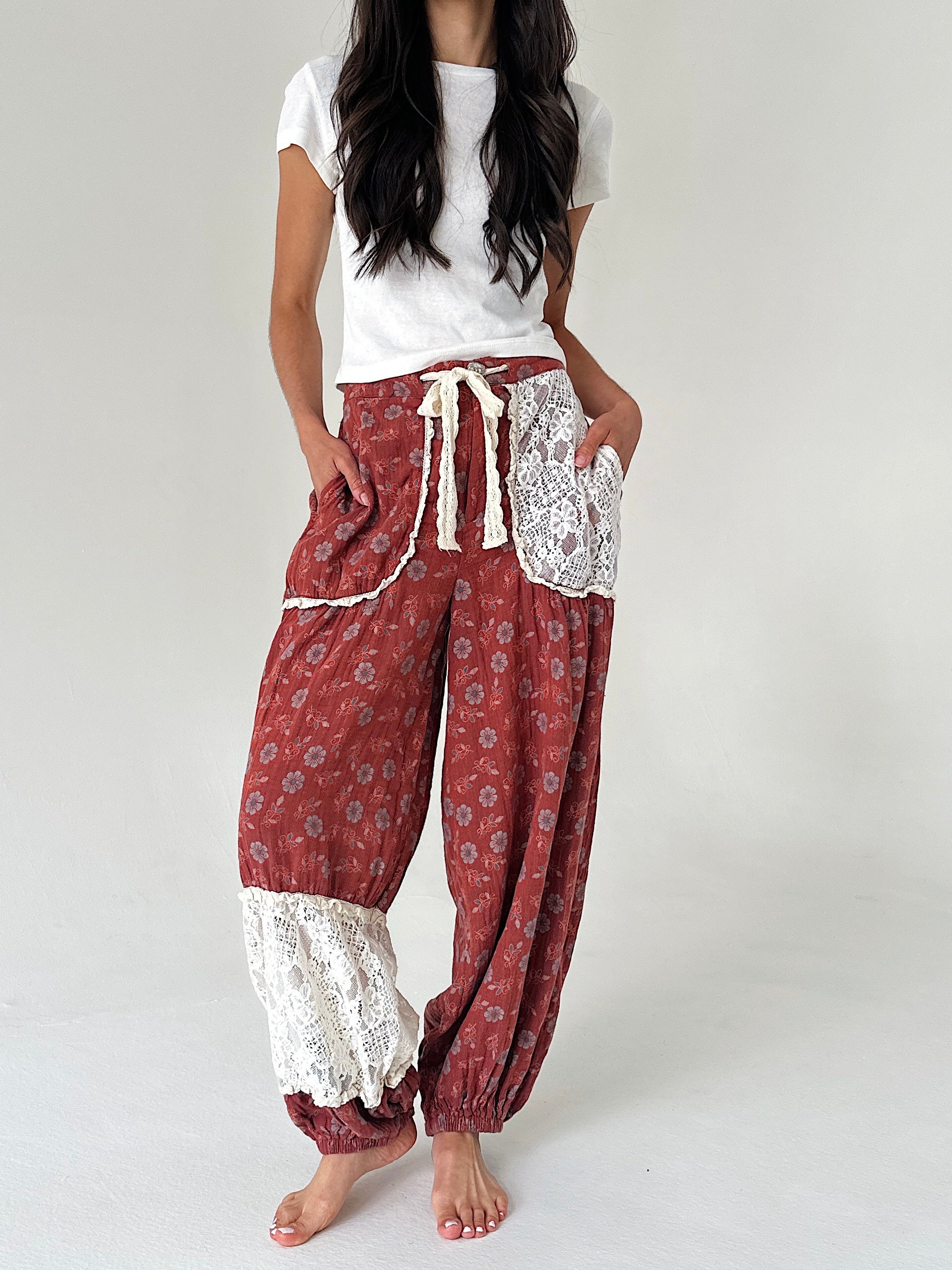 A model wearing loose, bohemian-style harem pants in a rust red floral print with white lace trim and a cream drawstring, paired with a white t-shirt, against a plain light gray background.