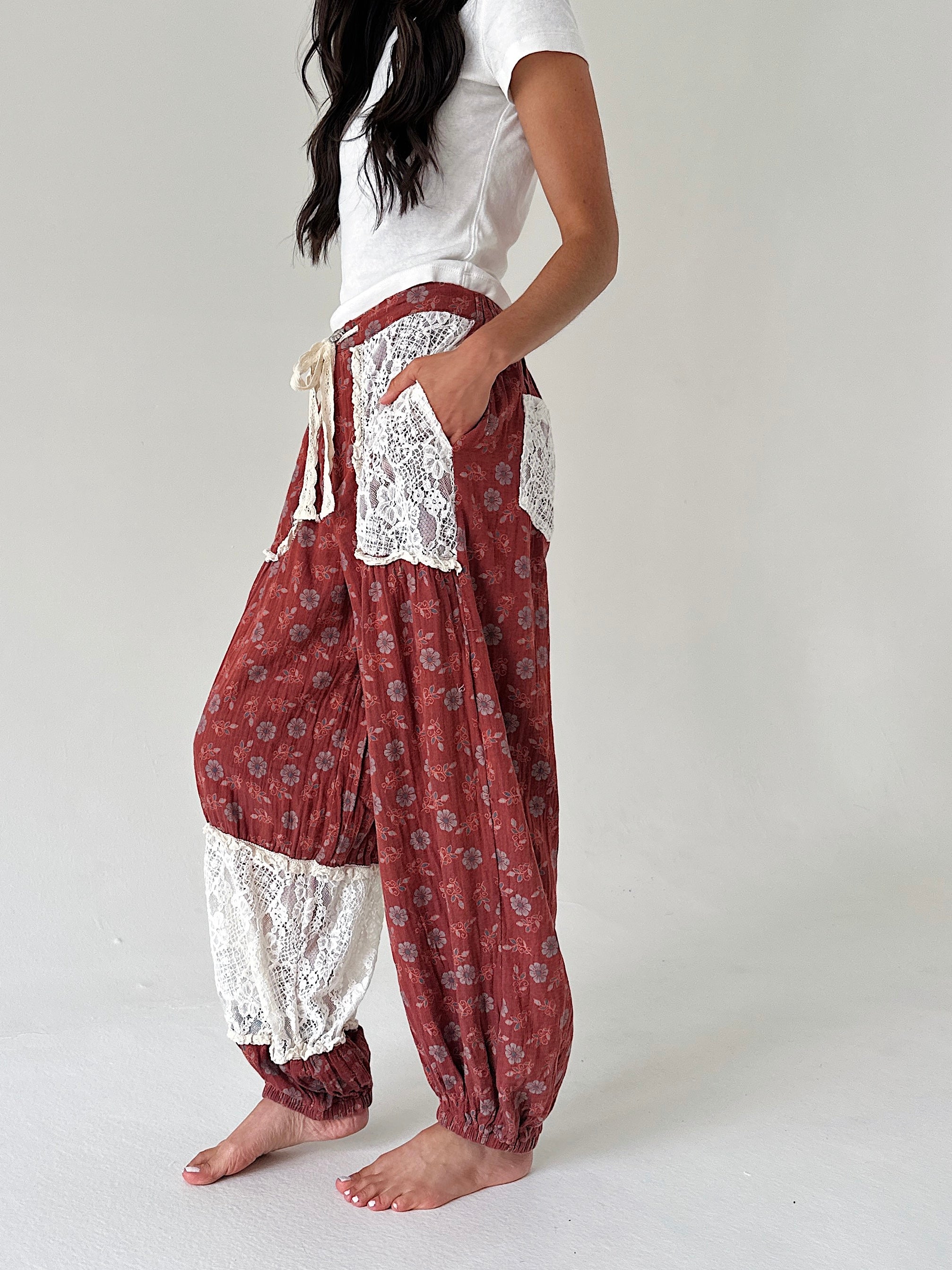 A person wearing loose, patterned harem-style pants in rust red with white lace trim, paired with a white t-shirt, standing barefoot against a plain light gray studio background.