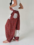 A person wearing loose, high-waisted harem-style pants in a rust red floral print with white lace pocket and lace lower-leg panels, paired with a white sleeveless top against a plain light gray studio background.