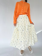 Person wearing an orange sweater and a white floral skirt on a plain background