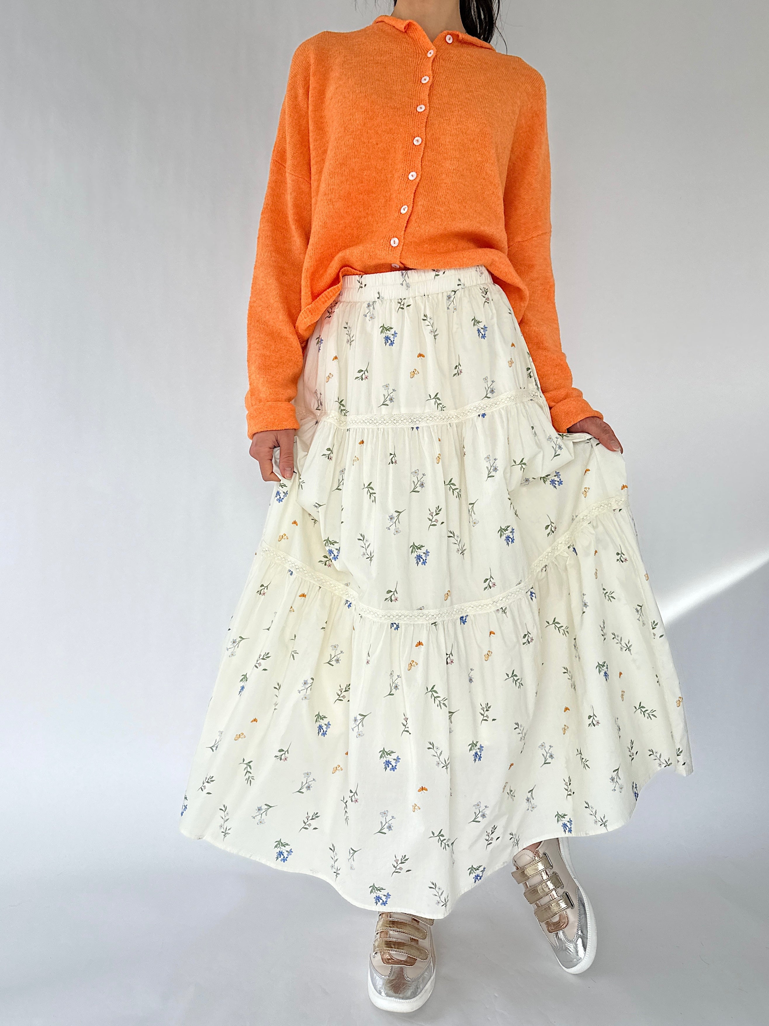 Person wearing an orange sweater and a white floral skirt on a plain background