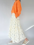 Person wearing an orange sweater and floral skirt on a plain background showing side pockets