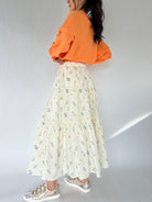 Person wearing an orange sweatshirt and a long floral skirt on a plain background