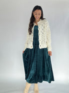 Woman wearing a white cardigan over a green velvet dress on a plain background