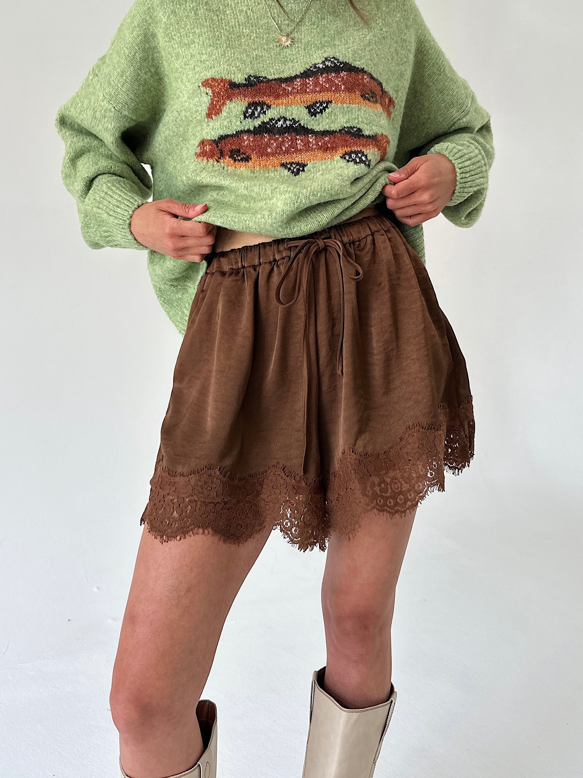 A person wearing a light green knit sweater with a fish pattern and brown shorts with a lace hem, paired with beige boots, against a plain light background.