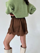 A person shown from the back wearing a light green fuzzy cropped sweater, a brown skirt with a lace hem, and off-white knee-high boots against a plain light gray background.