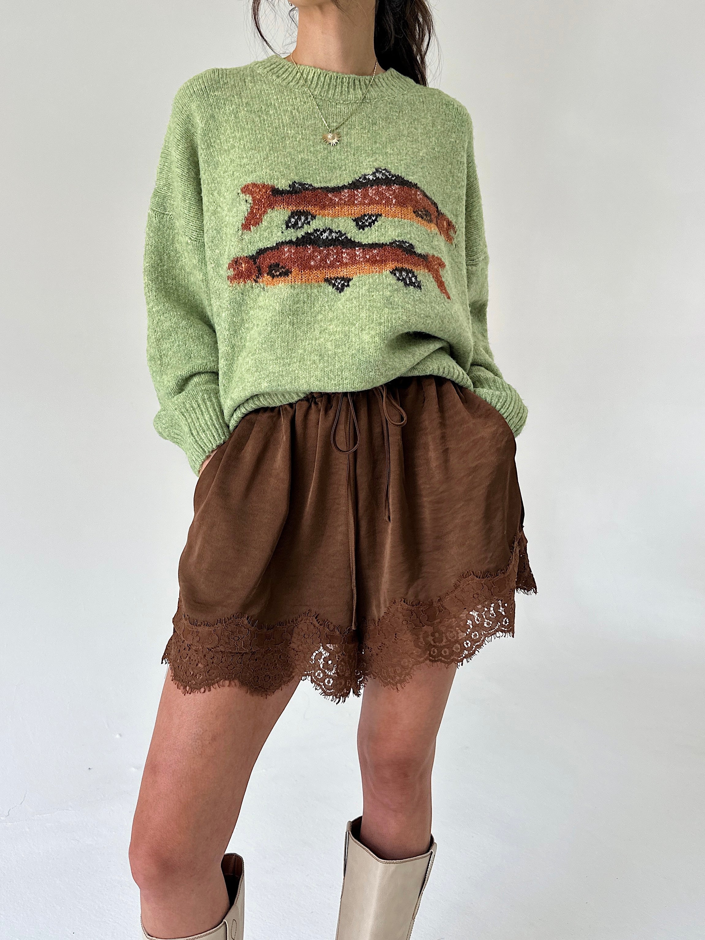 A model wearing a light green knit sweater with a brown and orange fish pattern on the chest, paired with brown shorts featuring a lace hem and beige boots, set against a plain white background.