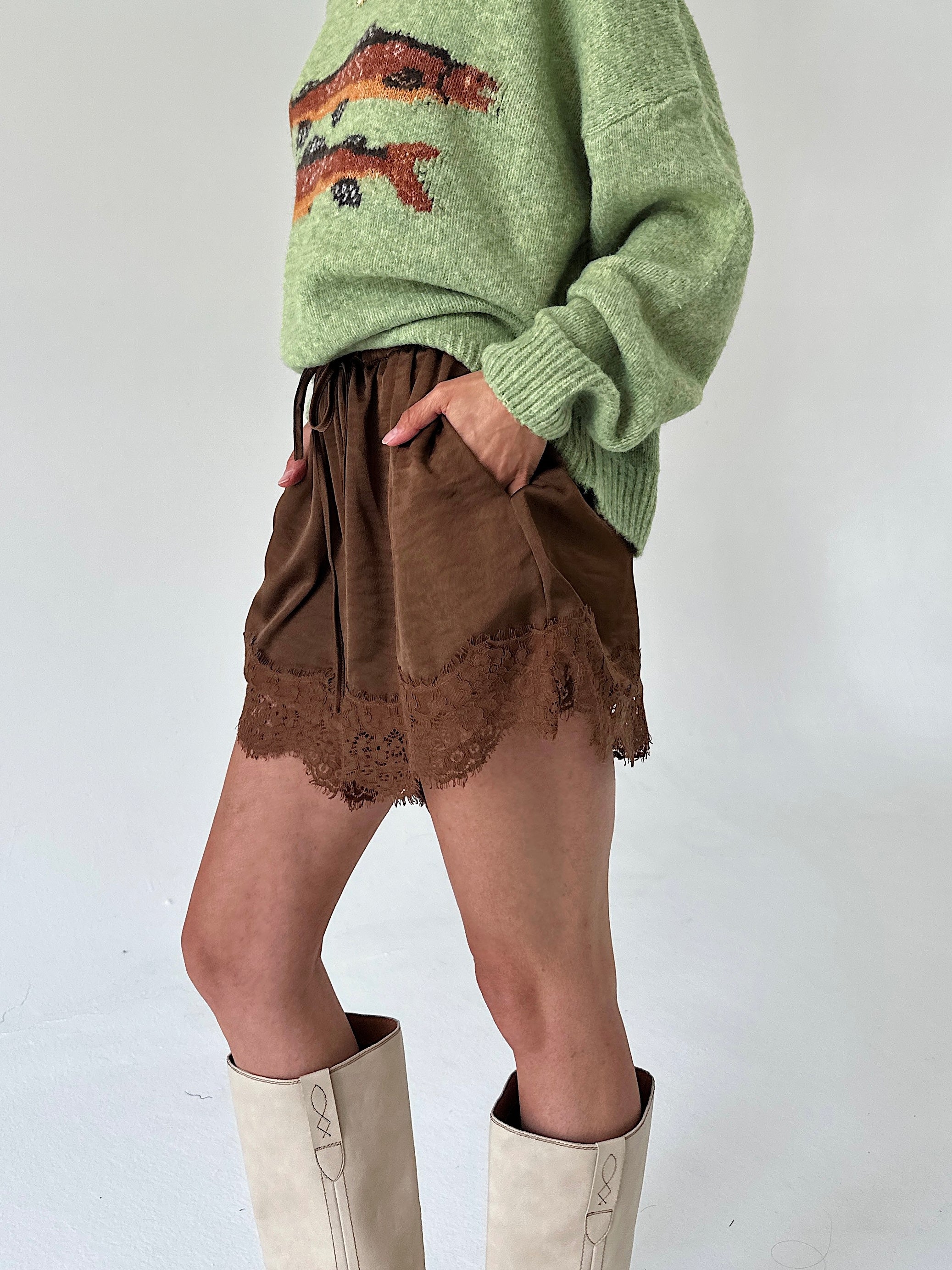 A person wearing brown satinshorts with a lace hem, paired with a light green sweater and off-white knee-high boots, shown against a plain light background.