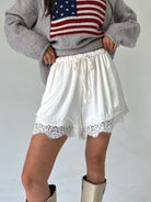 A person wearing white drawstring shorts with lace trim, paired with a gray sweater featuring an American flag graphic and beige boots, photographed against a plain light background.