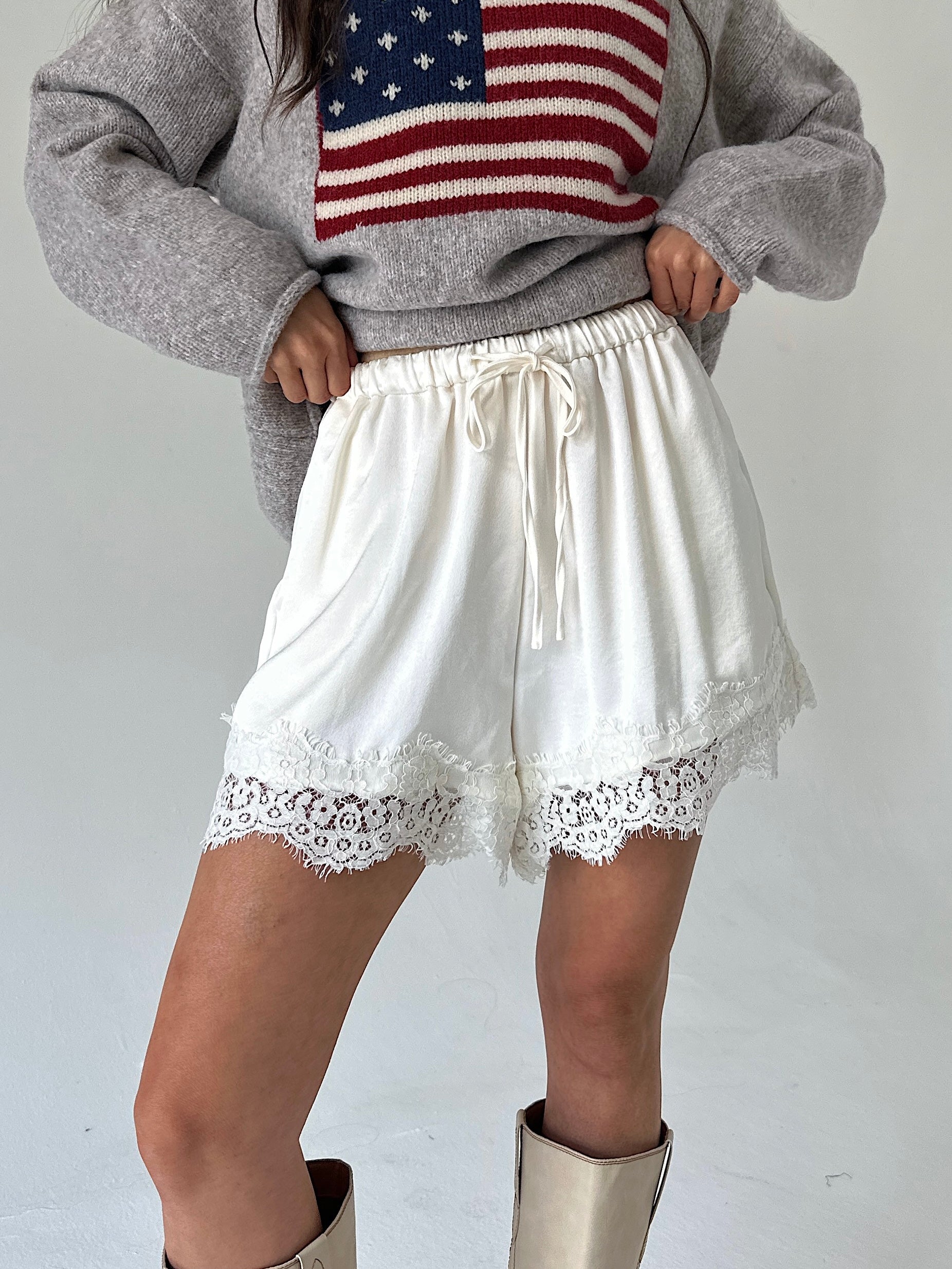 A person wearing white drawstring shorts with lace trim, paired with a gray sweater featuring an American flag graphic and beige boots, photographed against a plain light background.