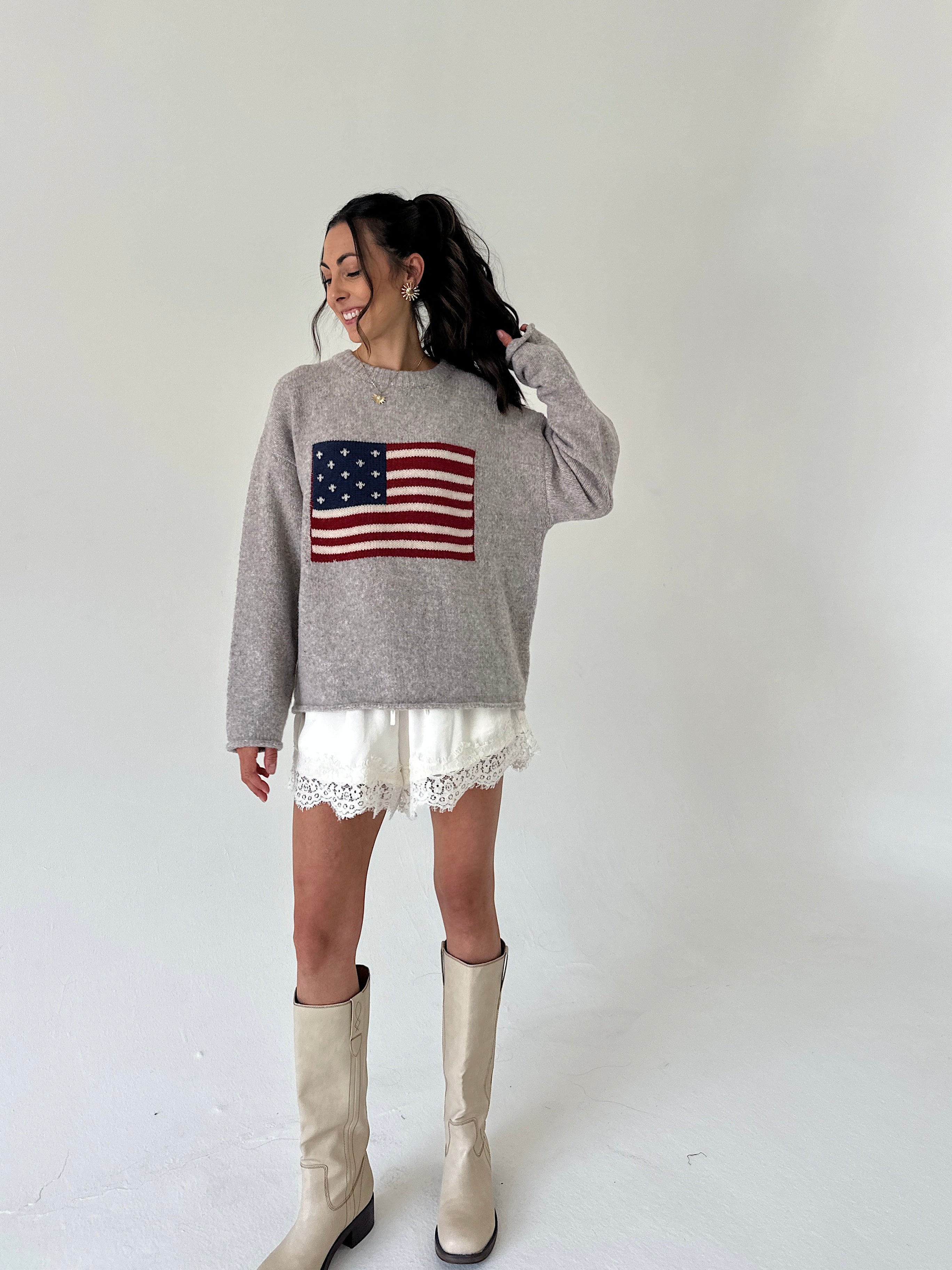 A woman modeling a heather gray crewneck sweater featuring a red, white, and blue American flag graphic on the front, paired with white lace-trimmed shorts and beige knee-high boots, set against a plain white studio background.