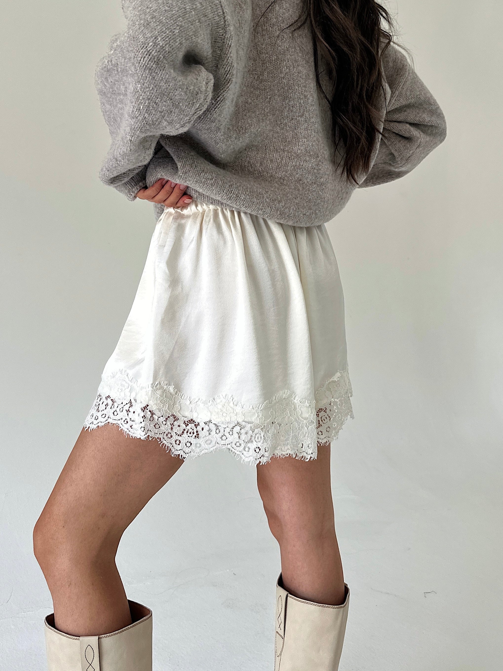 A person wearing white satin shorts with a lace hem, styled with a gray knit sweater and beige boots against a plain light background.