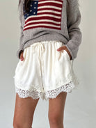 A person wearing white drawstring shorts with lace trim and a gray sweater featuring an American flag graphic, standing against a plain light background.