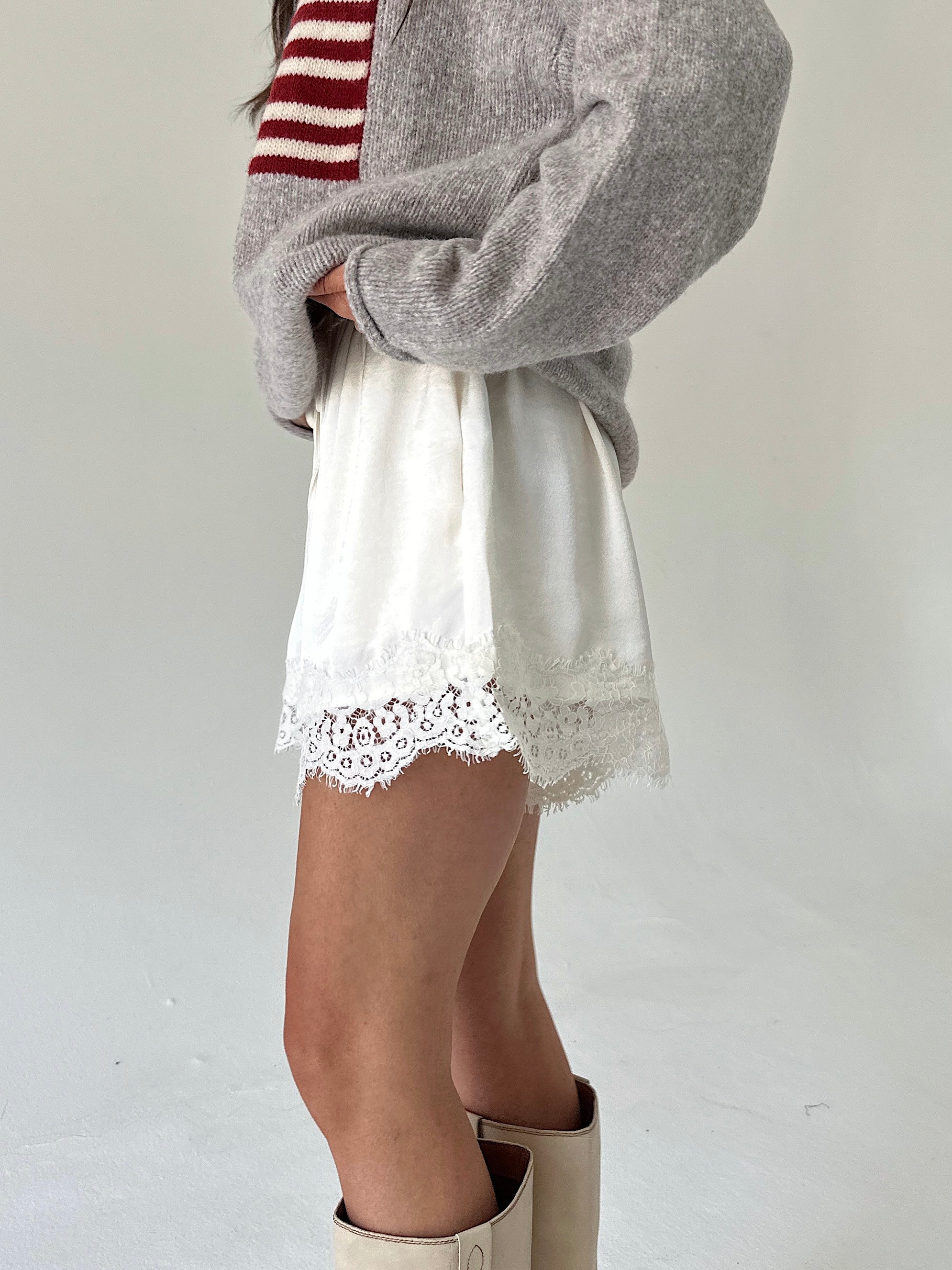 A person wearing a taupe knit sweater with a red and white stripe, white shorts with a lace hem, and beige boots, shown from the chest down against a plain light background.