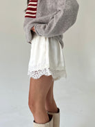 A person wearing a taupe knit sweater with a red and white stripe, white shorts with a lace hem, and beige boots, shown from the chest down against a plain light background.