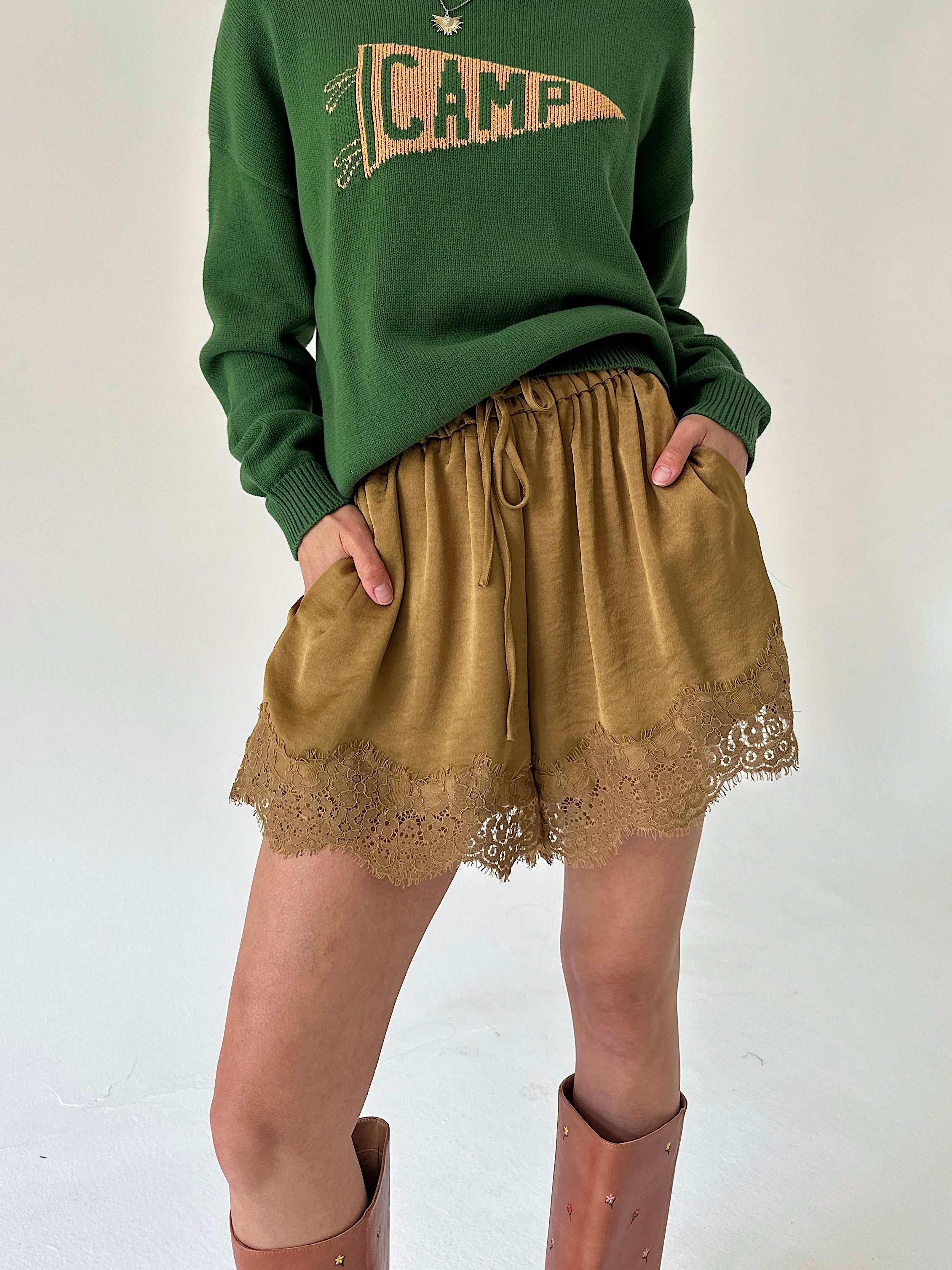 A person wearing a green crewneck sweater with a 'CAMP' pennant graphic and mustard satin shorts with a lace hem, paired with brown knee-high boots, against a plain off-white background.