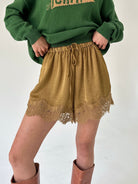 A person wearing olive green knit sweater and mustard drawstring shorts with lace trim, paired with brown boots, against a plain light background.