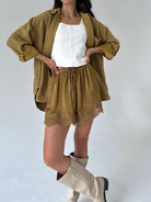 A model wearing a matching two-piece set consisting of a cropped, long-sleeve olive-brown shirt and matching drawstring shorts with lace trim, styled over a white tank top and cream-colored knee-high boots.