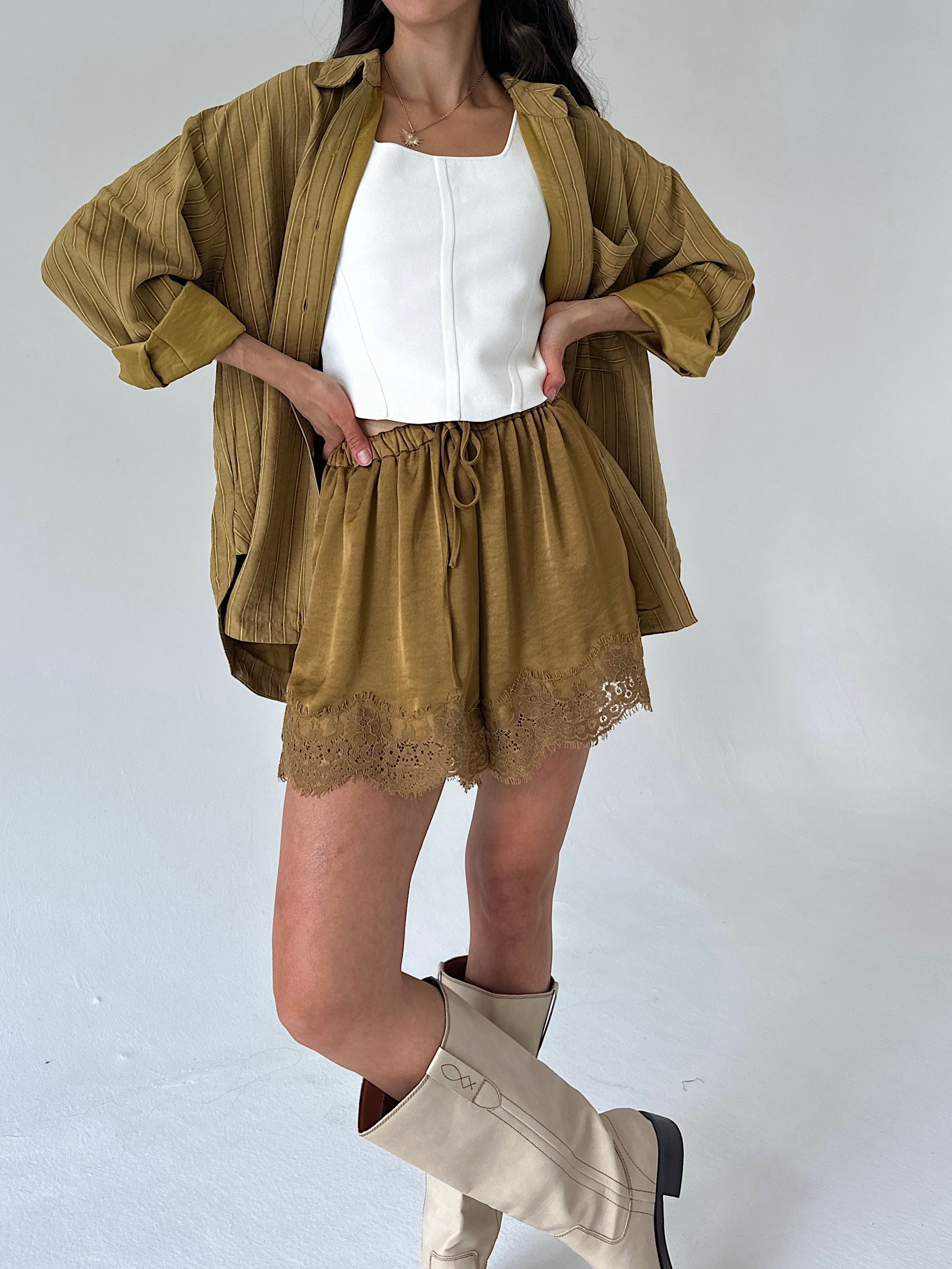 A model wearing a matching two-piece set consisting of a cropped, long-sleeve olive-brown shirt and matching drawstring shorts with lace trim, styled over a white tank top and cream-colored knee-high boots.