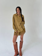 A woman modeling a matching mustard button-up shirt and lace shorts set, paired with brown leather knee-high boots, against a plain light gray studio background.