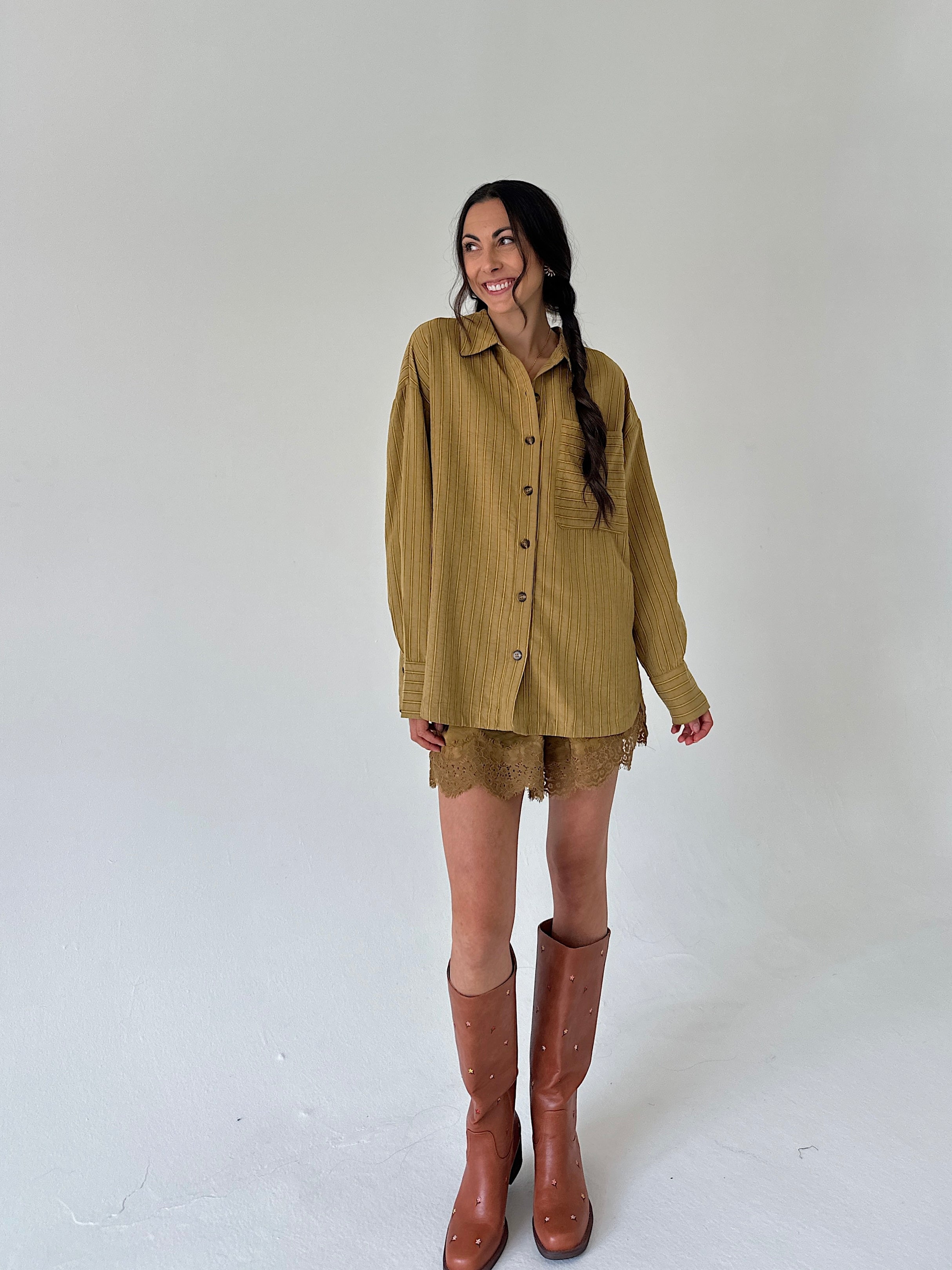 A woman modeling a loose olive-brown button-up shirt with matching lace shorts and brown knee-high boots, standing against a plain light gray studio background.