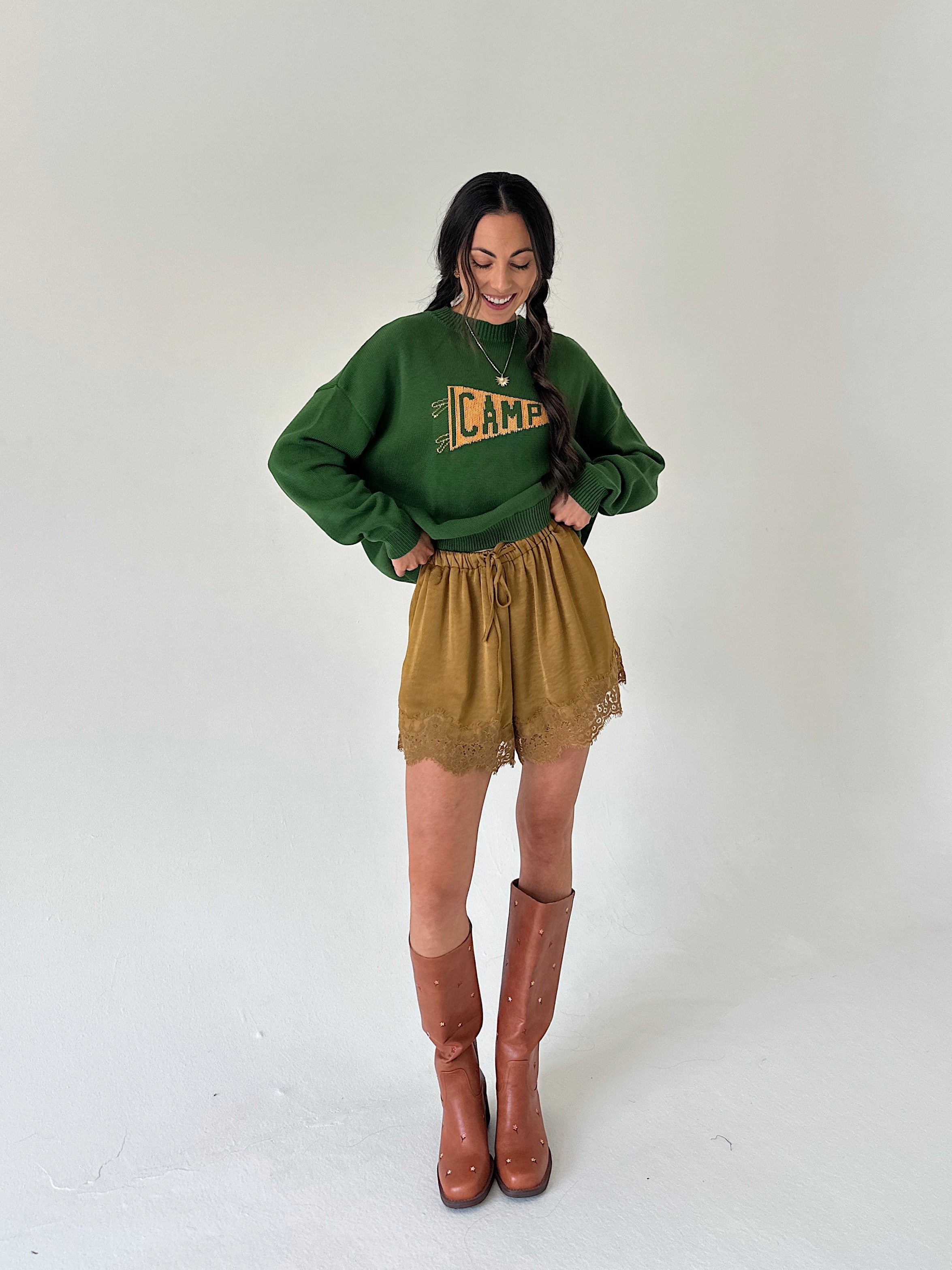 A woman modeling a dark green crewneck sweater with a yellow 'CAMP' graphic, paired with mustard lace-trim shorts and brown knee-high boots, against a plain light gray studio background.