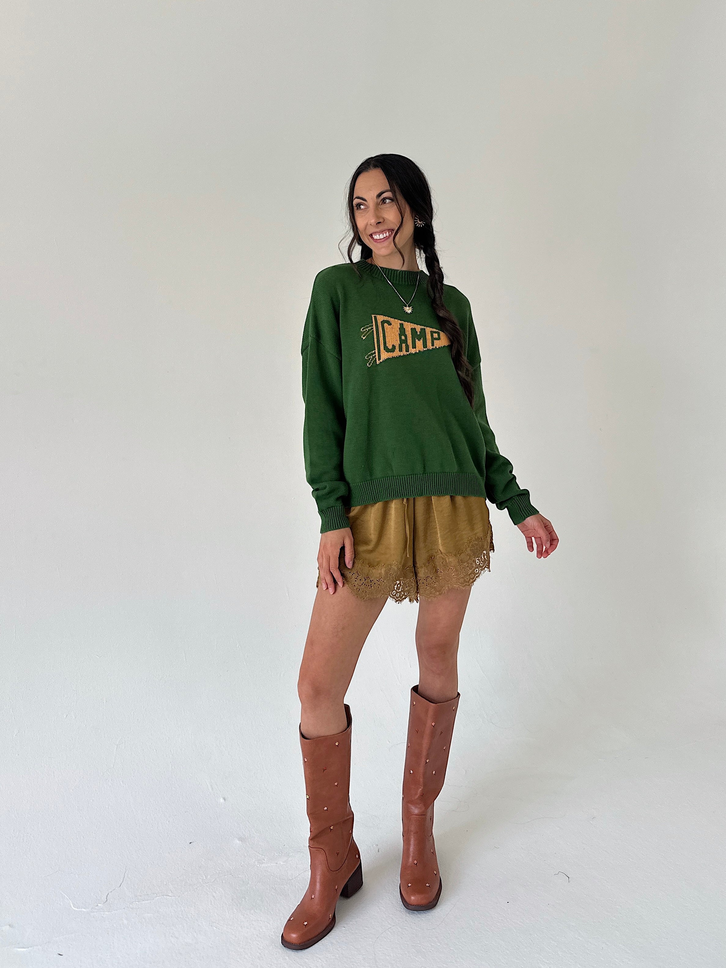 A woman modeling a dark green crewneck sweater with a distressed yellow 'CAMP' graphic, paired with mustard lace hem shorts and brown knee-high boots, against a plain off-white studio background.