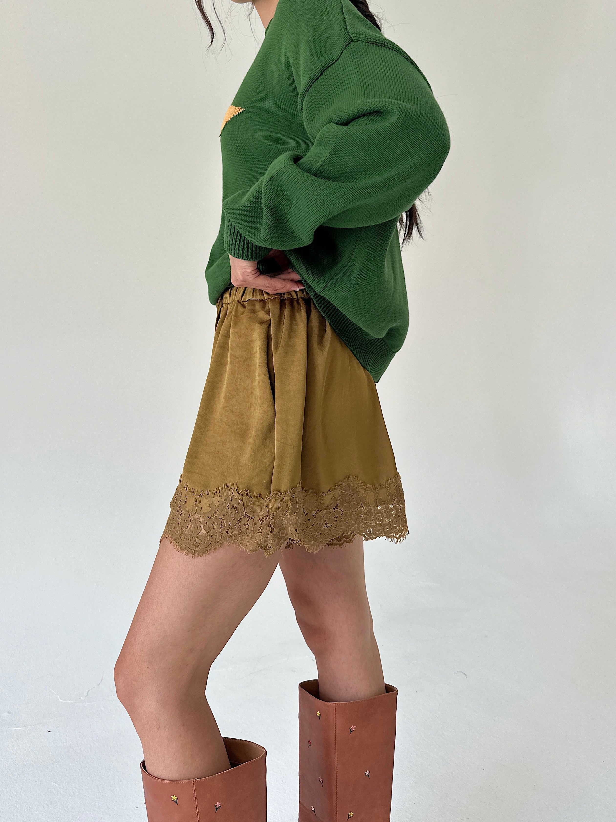 A person wearing an oversized green sweater, mustard shorts with a lace hem, and tall brown boots, photographed against a plain light background.