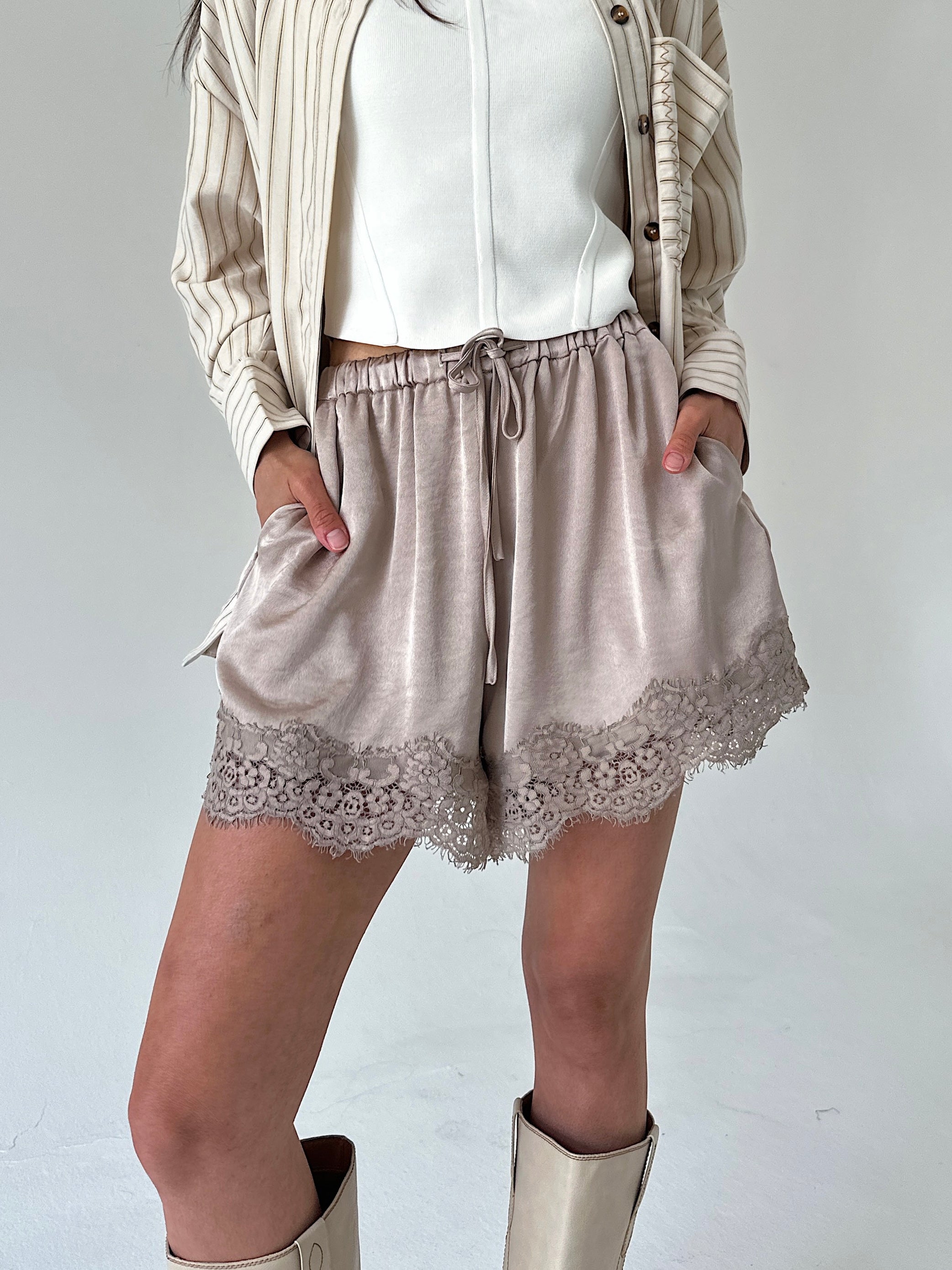 A person wearing light taupe satin shorts with a lace hem, styled with a white top, a beige striped cardigan, and beige boots against a plain light gray studio background.