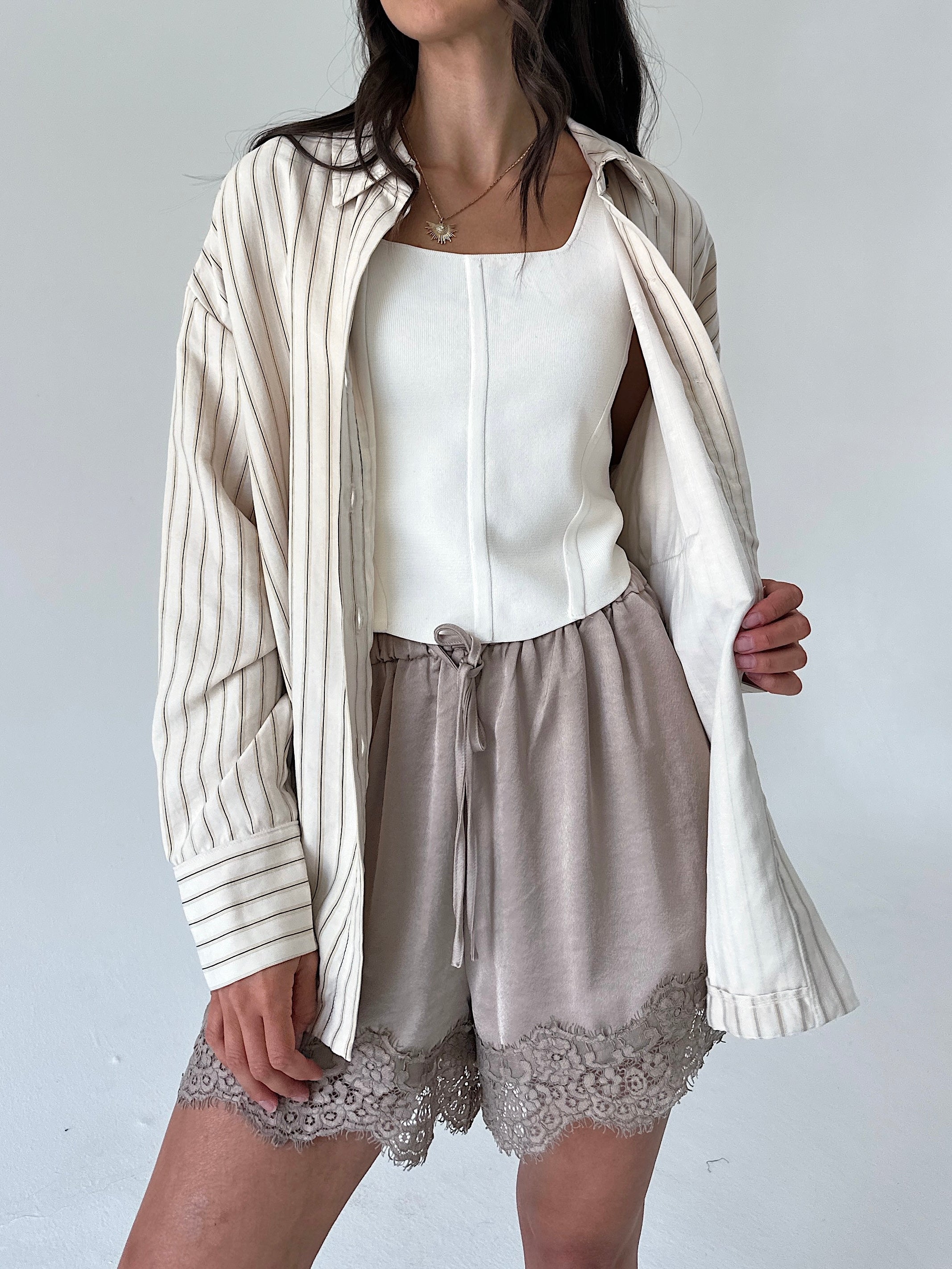 A model wearing a cream and beige vertically striped button-up shirt draped open over a white camisole, paired with taupe drawstring shorts featuring a lace hem.